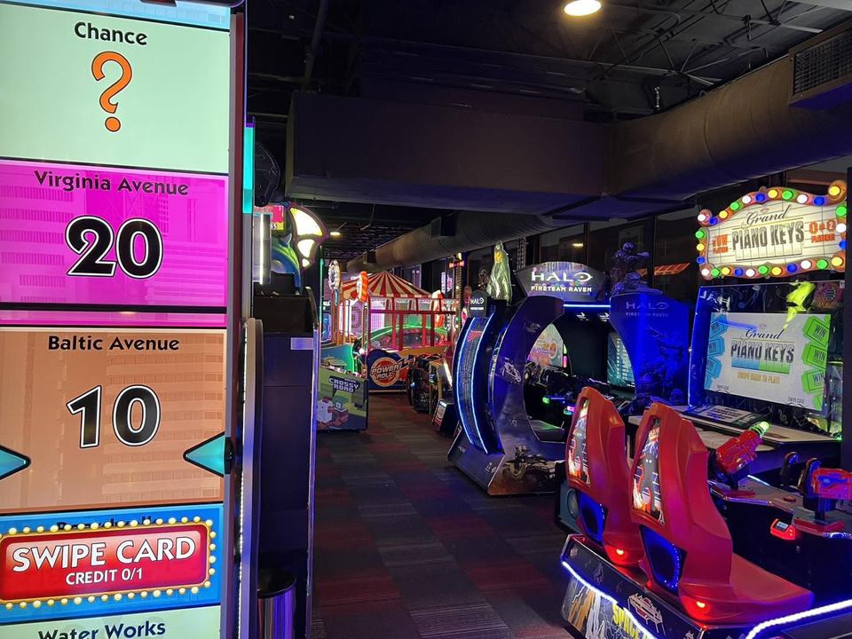 Down the arcade floor: a Monopoly ticket game with its Chance and Swipe Card panels, a racing cabinet and neon along the ceiling.