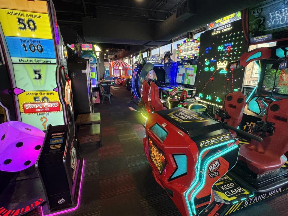 A row of neon arcade cabinets along the arcade aisle, a prize-ticket game in the foreground.