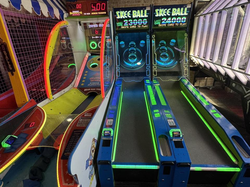 Two lit skee-ball lanes at the arcade, with neon and a racing cabinet beside them.