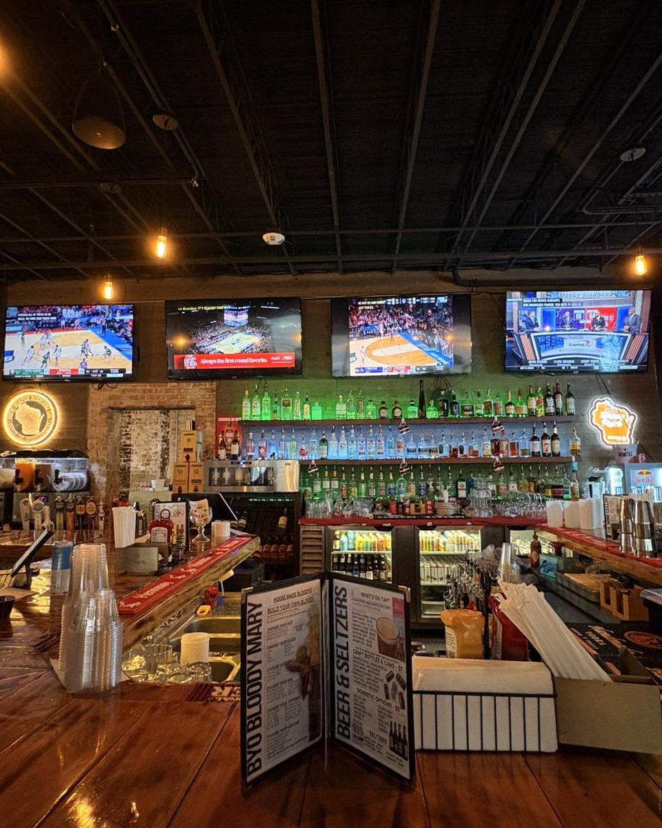 The empty bar under a dark ceiling: four TVs above a lit green back bar, a BYO Bloody Mary menu on the counter.