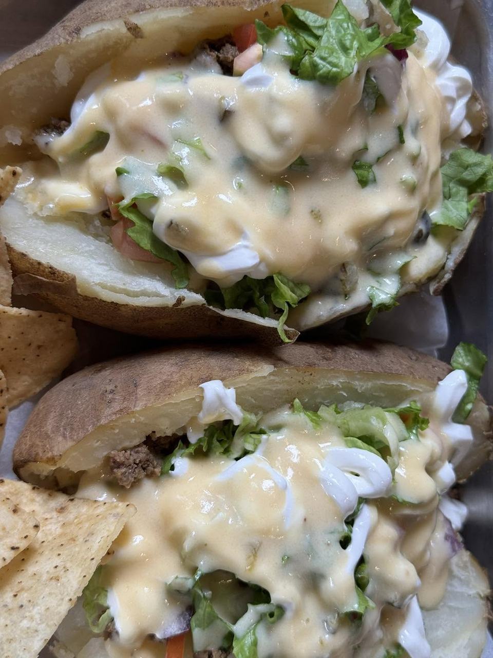 Two loaded baked potato halves topped with cheese sauce, lettuce, tomato and sour cream, beside tortilla chips.
