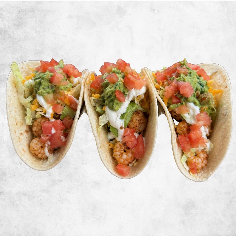 Three shrimp tacos in flour tortillas with guacamole, tomato and lettuce, on a white background.