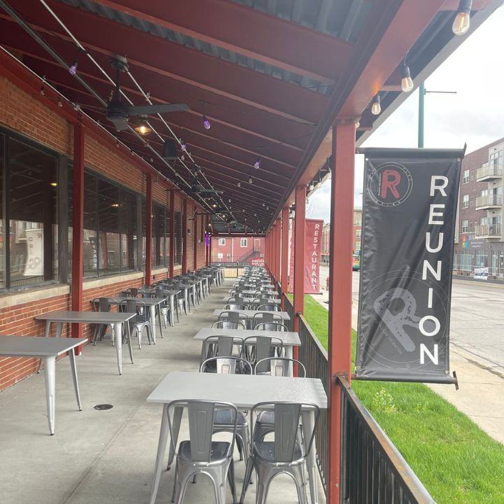 The covered brick patio in daylight: a row of metal tables and a black REUNION banner on a post.
