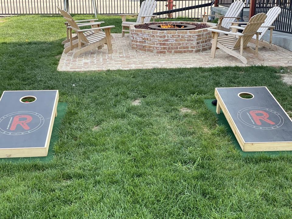 Two cornhole boards with the red R logo on the lawn, and a stone fire pit ringed with Adirondack chairs behind.