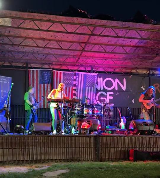 A band plays on the REUNION STAGE at night under pink and purple lights, seen from the lawn.
