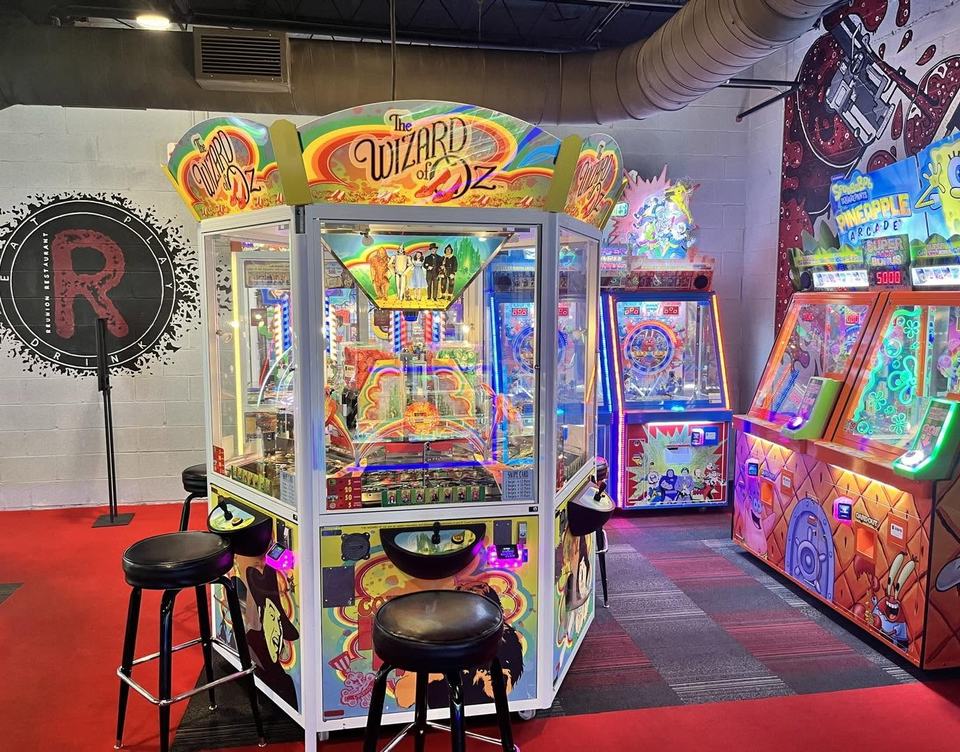 The round Reunion R logo painted large on the arcade wall with black spray splatter, beside a Wizard of Oz coin pusher, a red-lipped mural at top right.